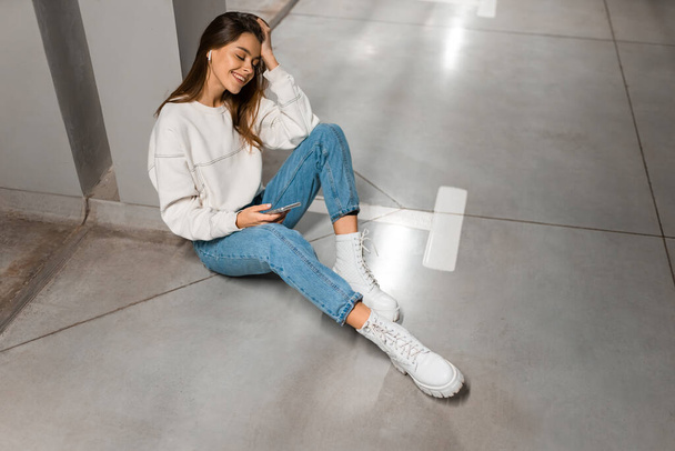 Fashion girl in de ondergrondse parking zit op de vloer en luistert naar muziek op draadloze koptelefoon vanaf smartphone - Foto, afbeelding