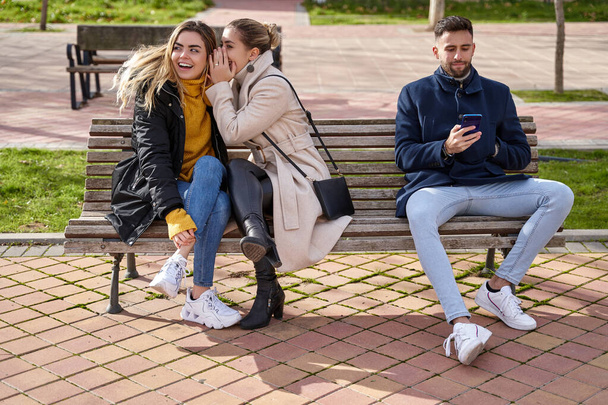 Dva krásní teenageři sdílejí tajemství, drby, zatímco se chlapec dívá na svůj mobil, sedí na lavičce v parku. - Fotografie, Obrázek