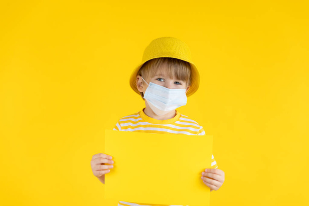 Happy child holding paper blank. Portrait of kid wearing protective mask indoor. Summer vacation during coronavirus COVID-19 pandemic concept. - Foto, Bild