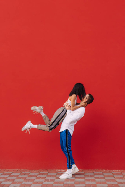 Passionate couple of stylish young man and woman in street clothes stand on a background of a red wall, a man holds a woman in his arms and passionately kisses. Valentine's Day. February 14th - Foto, afbeelding