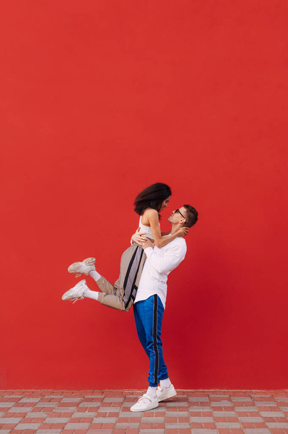 Cute kochający para w modnych ubrań casual stoi na tle czerwonej ściany i zabawy, mężczyzna podniósł kobietę w ramionach i wygląda z uśmiechem na twarzy. Walentynki. - Zdjęcie, obraz