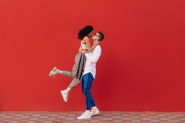 Stylowa młoda para zakochana bawiąca się na czerwonym tle ściany. Mężczyzna podniósł kobietę w ramionach i pocałował ją. Walentynki. Wielokulturowa miłość. - Zdjęcie, obraz