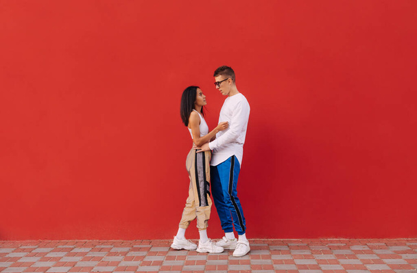 Módní bělošský pár a hispánská dívka objímají na červeném pozadí zdi s úsměvem na tvářích. Kokain muž a žena ve stylovém oblečení stojí na červeném pozadí. Multikulturní láska. - Fotografie, Obrázek