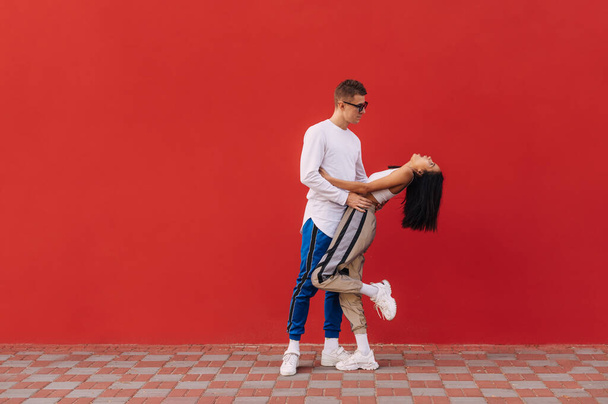 Stylowa para zakochana stojąca na ulicy na tle czerwonych ścian w modnych ubraniach, mężczyzna przytulający dziewczynę za talię. Walentynki. - Zdjęcie, obraz