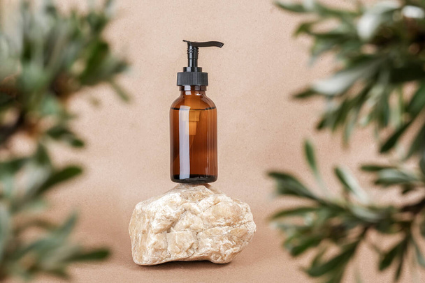Brown glass bottle with pump of cosmetic products on stone framed by green leaves of branches, beige background. Natural Organic Spa Cosmetic Beauty Concept. Front view Mock up. - Фото, зображення
