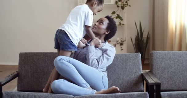 Afro american mom with daughter on couch hugging tenderly cuddling communicates talking joking smiling spending time together at home on weekend, motherhood and parenthood concept, ethnic family love - Video
