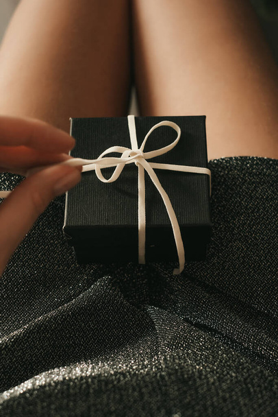 the hands of a girl in a silver dress holds a gift with a white ribbon in the red carpet. gift for new year, birthday, valentine's day. festive packaging. close-up - Foto, Imagem