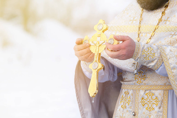 Τα χέρια ενός ιερέα βουτούν έναν ορθόδοξο χρυσό σταυρό στο ποτάμι. Πανηγύρι των Θεοφανείων της Ρωσίας. - Φωτογραφία, εικόνα