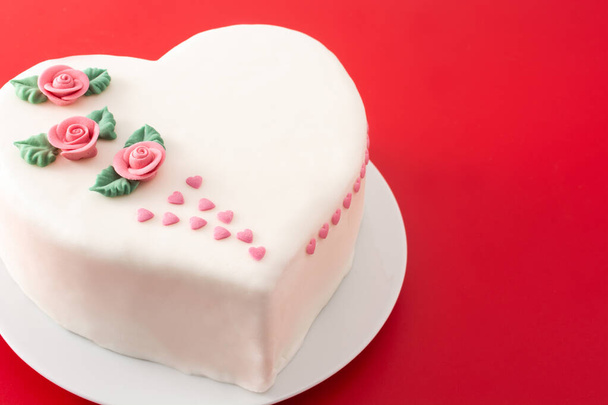 Καρδιά κέικ για την ημέρα του Αγίου Βαλεντίνου, Ημέρα της μητέρας, ή γενέθλια, διακοσμημένα με τριαντάφυλλα και ροζ καρδιές ζάχαρη στο κόκκινο φόντο - Φωτογραφία, εικόνα
