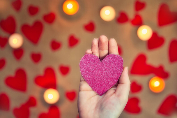 Mão feminina segurando um coração rosa brilhante com vela romântica e fundo de corações vermelhos para o dia dos namorados ou conceito romântico - Foto, Imagem
