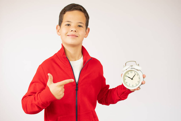 childhood, fashion and people concept - portrait of happy smiling boy in red sweater with alarm clock over white background - Photo, Image