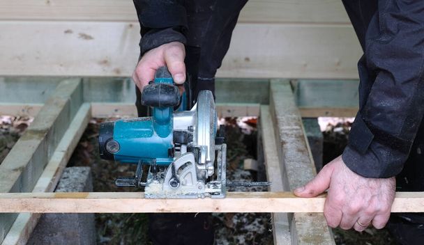 egy építőipari vállalkozó kézi körfűrésszel vágja fel a deszkákat, miközben házat épít - Fotó, kép