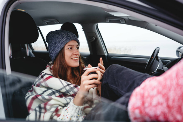 Handsome girl traveler wearing boho scarf rest in car and looking away winter landscape. Young woman traveler sitting at car and get warm cup of tea - 写真・画像