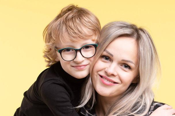 Portrait of a happy young woman with her son, faces touching each other. The child with glasses hugged mom with love. Caucasian. Emotional faces. Yellow studio background, horizontal. - 写真・画像