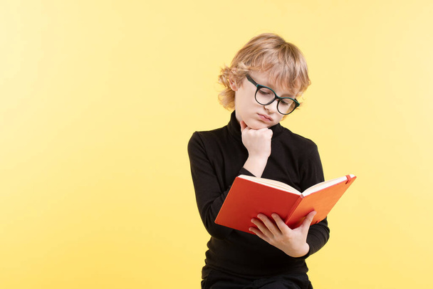 Blond blanke nerdschooljongen in een zwarte trui, met een bril op, denkend aan een open notitieboekje. Kind verveelt zich met het lezen van de tekst tijdens het doen van huiswerk. Geïsoleerd op gele studio achtergrond.  - Foto, afbeelding