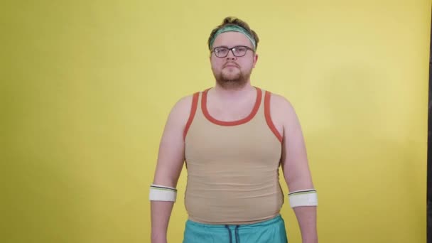 Funny fat man in sportswear with dumbbells in his hands. Overweight man goes in for sports. Yellow background isolate - Filmagem, Vídeo