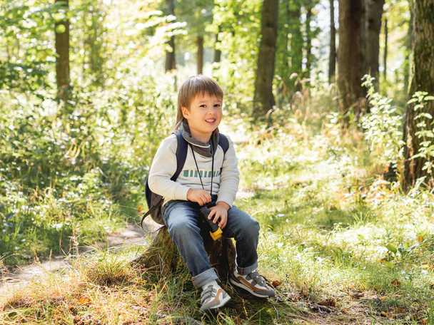 Little explorer on hike in forest. Boy with binoculars sits on stump. Outdoor leisure activity for children. Summer journey for young tourist. - Fotó, kép