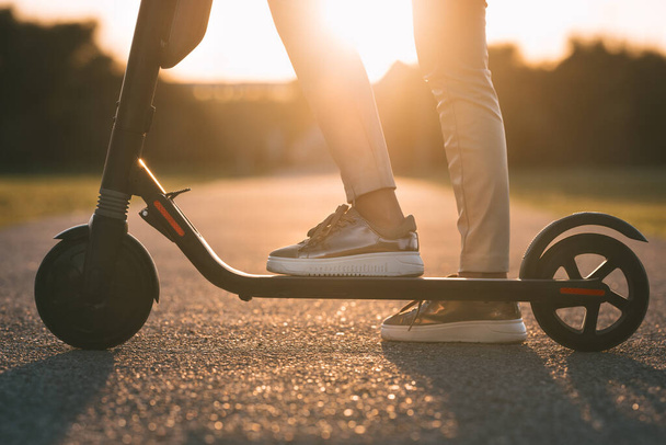 Close up of woman riding electric kick scooter at sunset. Electric urban transportation - 写真・画像