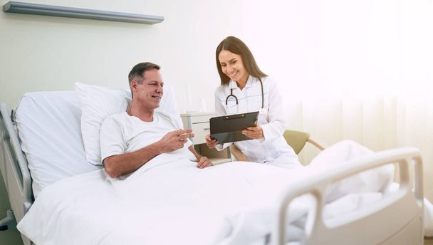 bello maturo paziente uomo è sdraiato su il clinica letto e ha un conversazione con un fiducioso giovane donna medico in un bianco cappotto - Foto, immagini