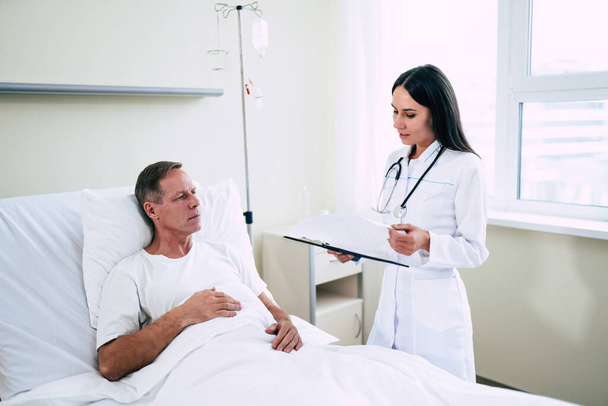 Handsome mature patient man is lying on the clinic bed and has a conversation with a confident young woman doctor in a white coat - Photo, image