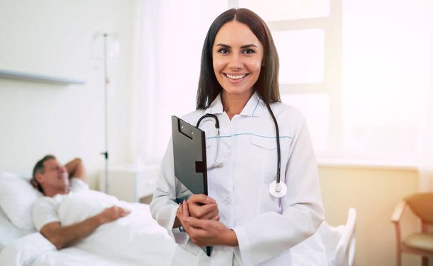 Close up photo of a smiling confident cute woman doctor in a coat with a stethoscope is looking on the camera with the patient in bed in the background  - Photo, Image