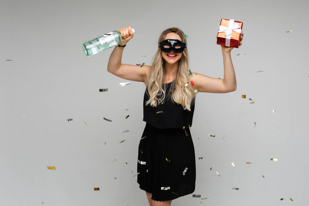 happy young woman in short balck dress and mask celebrates new year with a bottle and a present - Фото, зображення