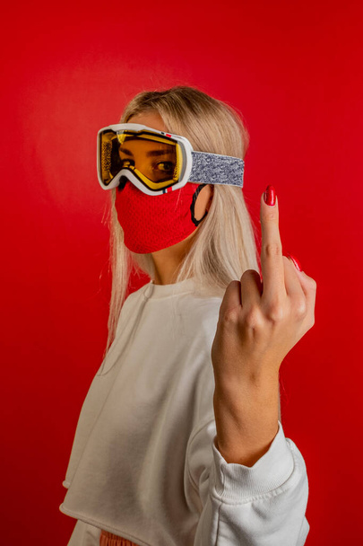 Close-up girl skier wearing a red protective mask and glasses doing middle finger gesture. A snowboarder on red background showing middle finger doing fuck you bad expression. Covid19 coronavirus and the ski resort pandemic - Photo, image