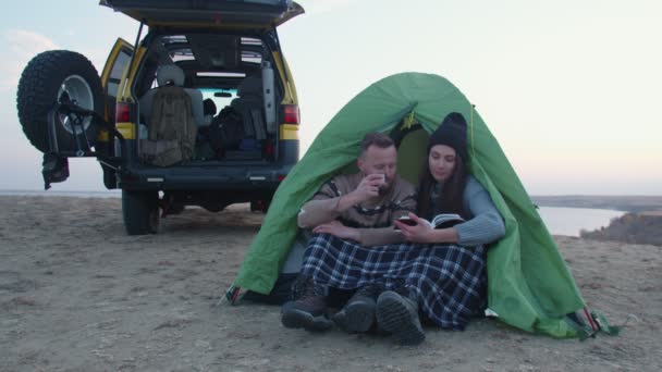 Young man and woman sits near the tent and the car, drinks a hot tea and read the book in the mountain - Footage, Video