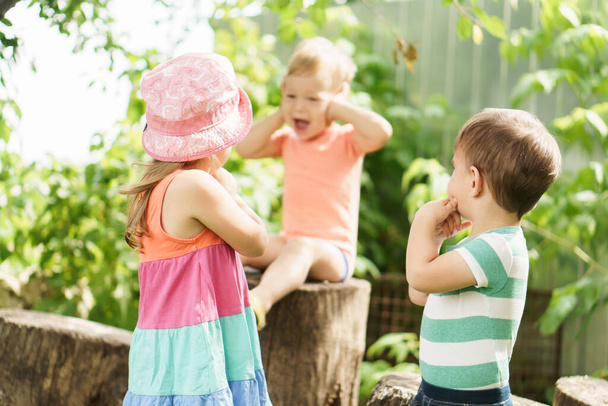 Three children of same age show different emotions. Kids in green garden play game deaf blind dumb outdoors in summer. sister and brothers show gesture of silence, hold finger to their lips, cover - Photo, image