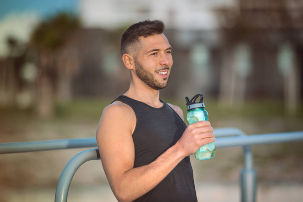 Joven hombre de fitness beber agua con limón de la botella reutilizable durante un entrenamiento. Beber mientras se hace ejercicio. Deshidratación durante el entrenamiento - Foto, Imagen