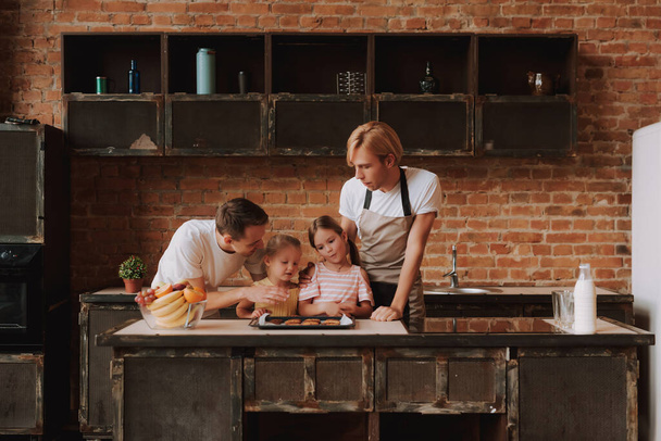 Gay couple with their adopted cute daughters cooking on kitchen. Lgbt family at home. Portrait of two handsome men and their two little daughters eating cookies. - Foto, immagini