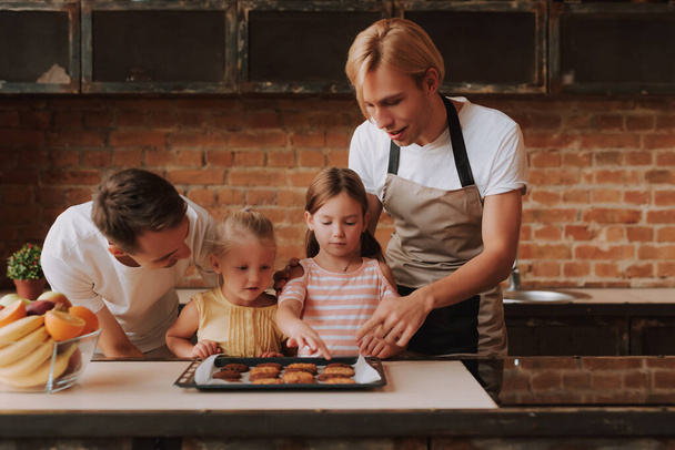 Gay couple with their adopted cute daughters cooking on kitchen. Lgbt family at home. Portrait of two handsome men and their two little daughters eating cookies. - Photo, Image
