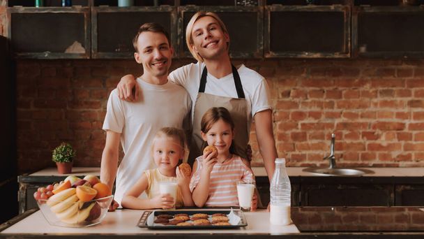 Meleg pár az örökbefogadott aranyos lányaikkal főznek a konyhában. A család nem otthon van. Két jóképű férfi és két kislányuk portréja, akik sütit esznek és tejet isznak.. - Fotó, kép