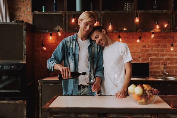 Loving gay couple at home. Two handsome men hugging and kissing on kitchen. Romantic atmosphere with bottle of red wine. LGBT concept. - 写真・画像