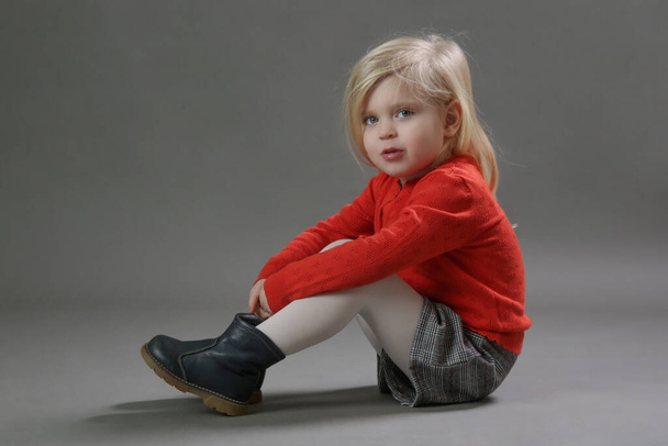 Retrato de estudio de moda de adorable niña de 3 años en suéter rojo abotinado y pantalones cortos a cuadros sobre fondo gris. - Foto, imagen