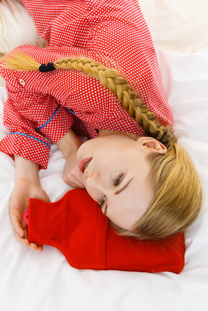 Woman lying on bed sleeping with warm red hot water bottle. Fever, flu treatment objects concept. - Zdjęcie, obraz