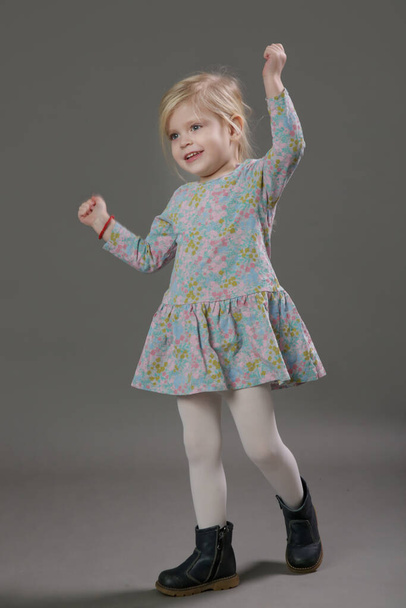 Fashion studio portrait of adorable 3 years old blonde girl in floral pattern dress on grey background. - Fotó, kép