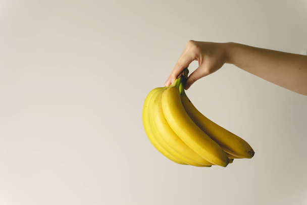 Κοντινό πλάνο στο χέρι του αγνώστου καυκάσιος γυναίκα κρατώντας μπανάνες μπροστά από λευκό τοίχο - υγιεινά βιολογικά τρόφιμα φρέσκα φρούτα έννοια αντίγραφο χώρο - Φωτογραφία, εικόνα