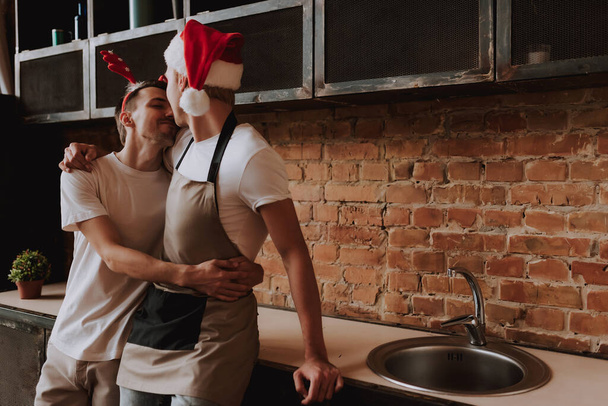 Kochająca para gejów w domu. Dwóch przystojniaków przytulających i całujących się w kuchni w kapeluszu Świętego Mikołaja. Boże Narodzenie. Koncepcja LGBT. - Zdjęcie, obraz