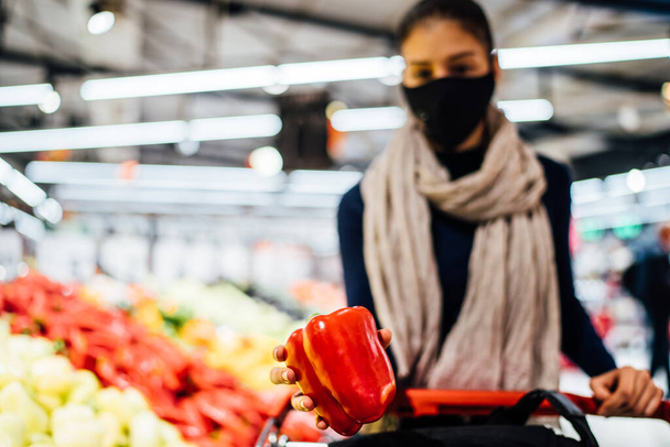 Young woman wearing protective face mask shopping in a supermarket,buying organic produce and ingredients.Eating healthy food during coronavirus COVID - 19 pandemic.Healthy bell pepper.Grocery shop - Photo, image