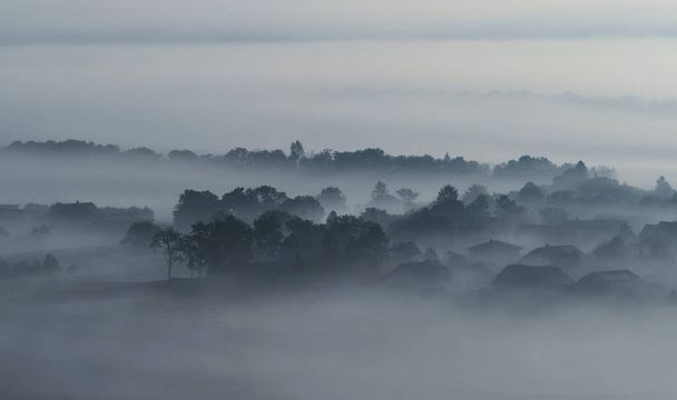 Edificios de la aldea casas y árboles en las nubes de niebla densas bajas neblina de la mañana malhumorado en Gemeinlebarn Baja Austria - Foto, Imagen