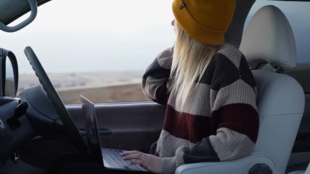 Young female freelancer in yellow hat sits in car and works with notebook outdoors - Footage, Video