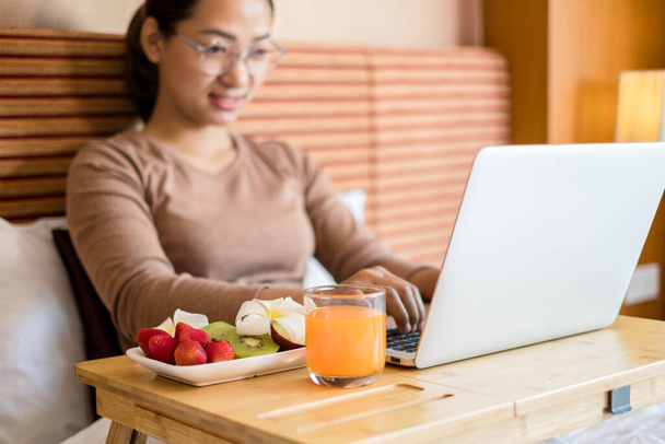Focus on fruit. young Asian women smiling happily at freelance work, working on a notebook while relaxing in bed with snacks and fruit in a hotel room. Vacation and relaxation. - Photo, Image