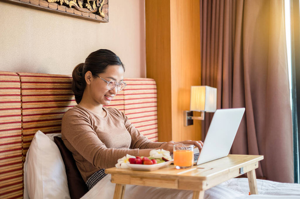 Genç Asyalı kadınlar serbest çalışmada mutlu bir şekilde gülümserken otel odasında aperatifler ve meyvelerle yatakta dinlenirken bir defter üzerinde çalışıyorlar. Tatil ve rahatlama. - Fotoğraf, Görsel