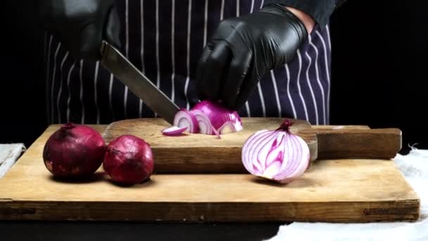 chef en guantes de látex negro corta cebollas rojas en una tabla de cortar de madera, negro - Imágenes, Vídeo