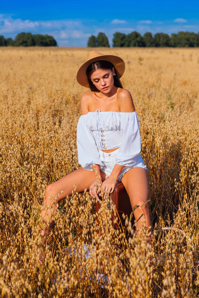 Rural Countryside Scene. Fashion portrait of a young beautiful woman with long hair dressed in white blouse and straw hat sitting on the old vintage suitcase at golden oat field. - Foto, Imagen
