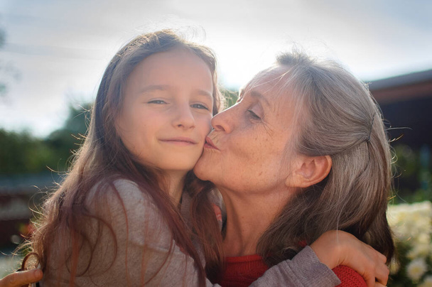 Abuela mayor con el pelo gris vistiendo suéter rojo con su nieta pequeña se abrazan en el jardín y durante el día soleado al aire libre, día de las madres - Foto, imagen