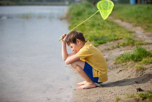 Un petit garçon avec un filet à papillons est assis près du lac et regarde l'eau. - Photo, image