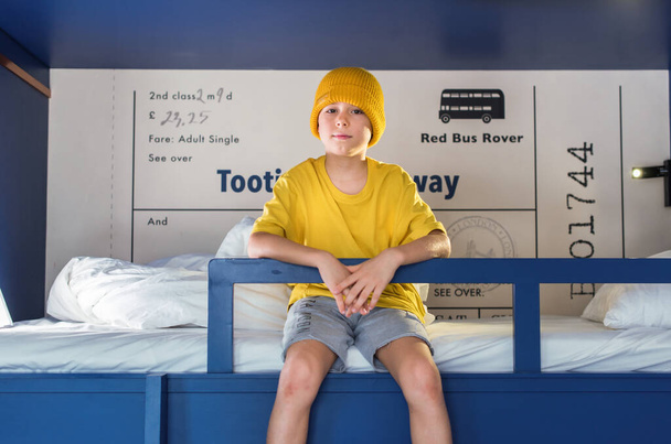 Boy in yellow t-shirt and hat is spending time sitting gown on a blue two-tier bunk bed at home. - Foto, Bild