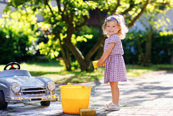 Menina bonito lindo bebê lavando grande carro de brinquedo velho no jardim de verão, ao ar livre. Feliz saudável pequena criança limpando carro com água e sabão, se divertindo com espirrar e brincar com esponja. - Foto, Imagem